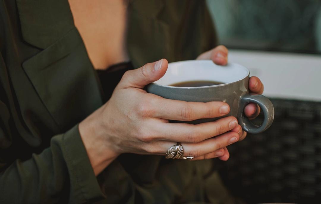 Les mains d'une femme en veste verte entourent une tasse de café grise ; à son doigt, une bague argentée en forme de feuille.