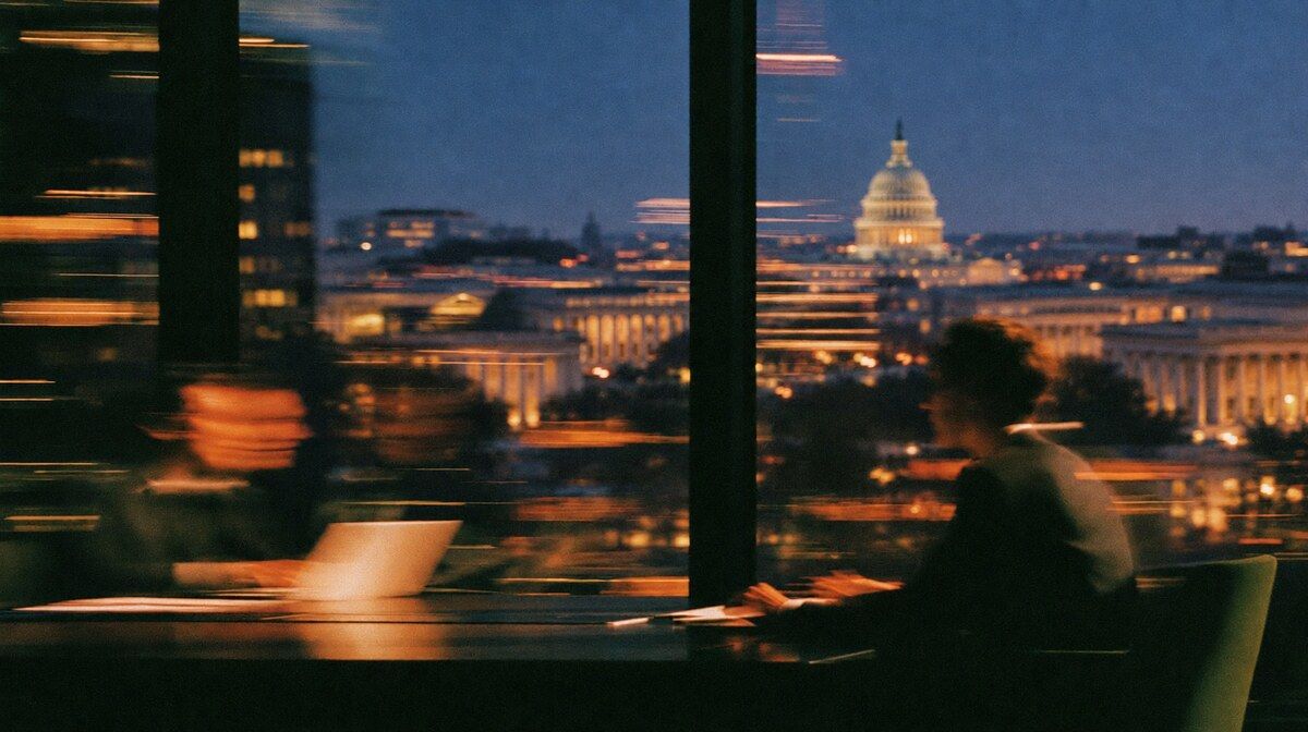People working on laptops with the U.S. Capitol visible through a window at dusk.