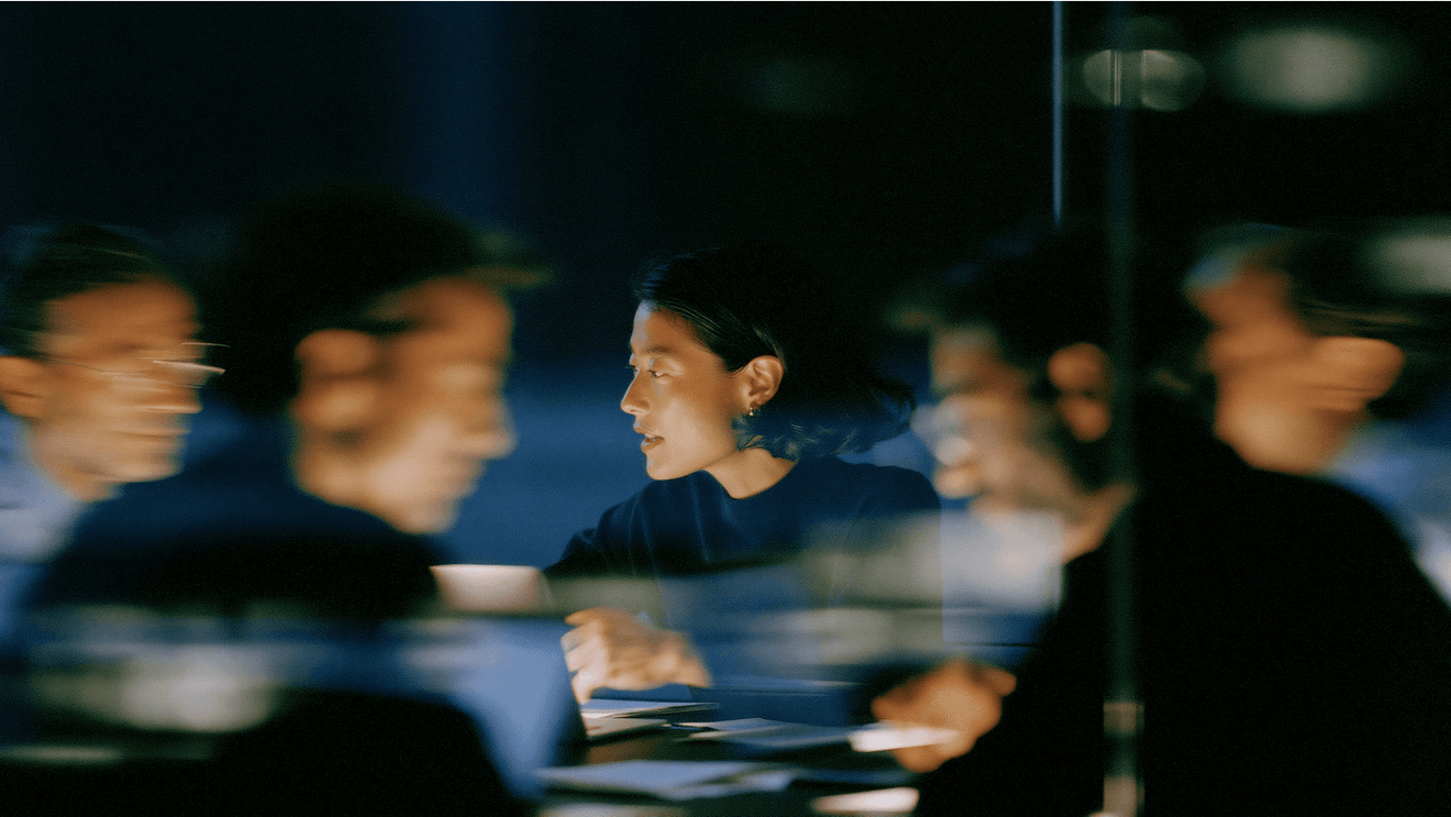 A woman works at a laptop in a dimly lit room, her face in sharp focus while motion-blurred colleagues stream past her on either side.