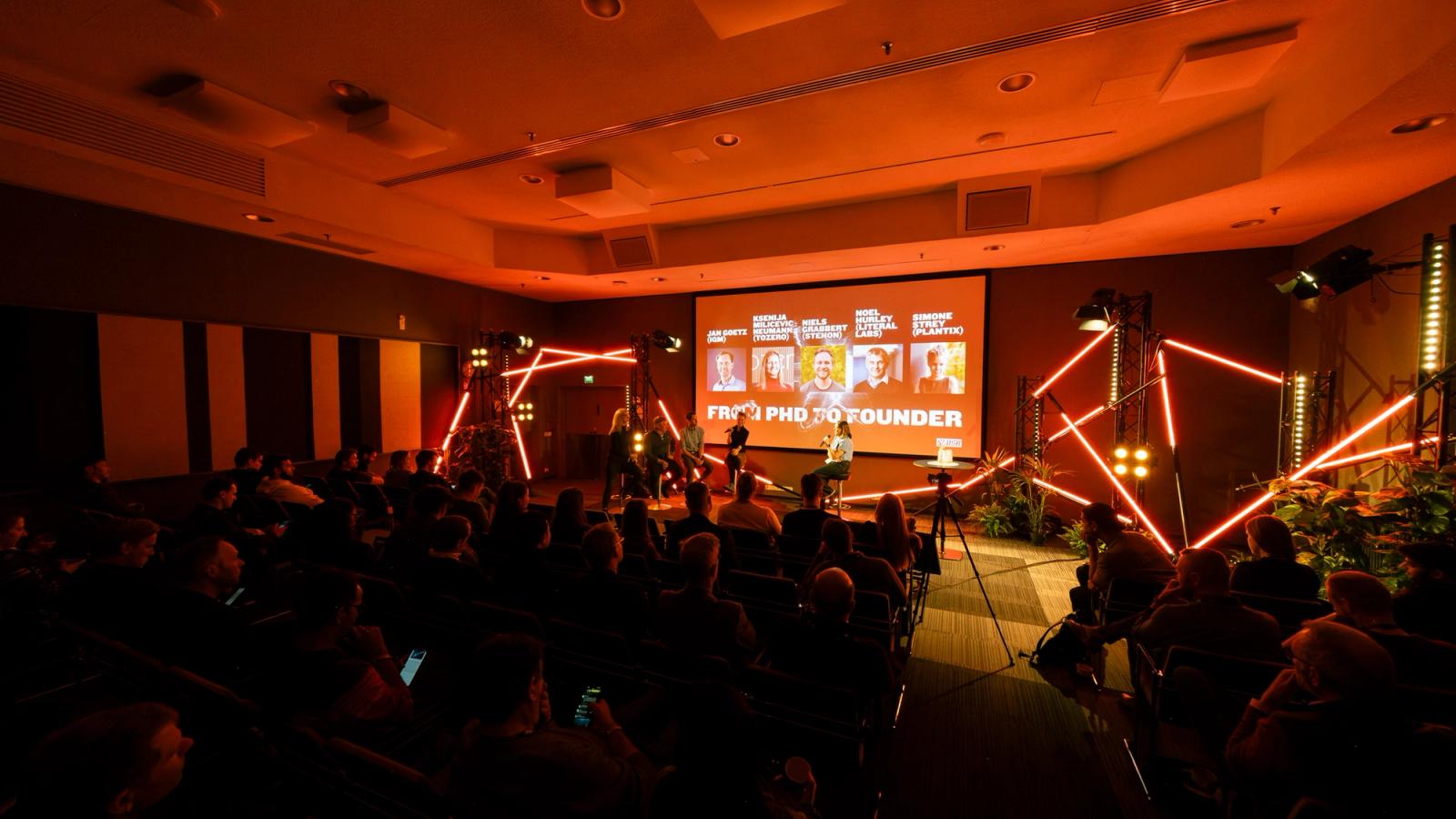 A seated audience in a dark room watches a panel on a small stage lit by red neon tubes, under a screen that reads From PhD to Founder.
