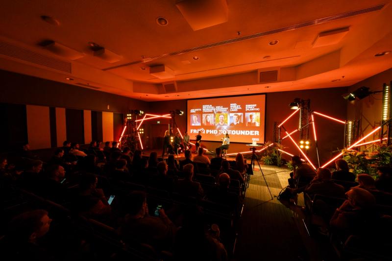 A seated audience in a dark room watches a panel on a small stage lit by red neon tubes, under a screen that reads From PhD to Founder.