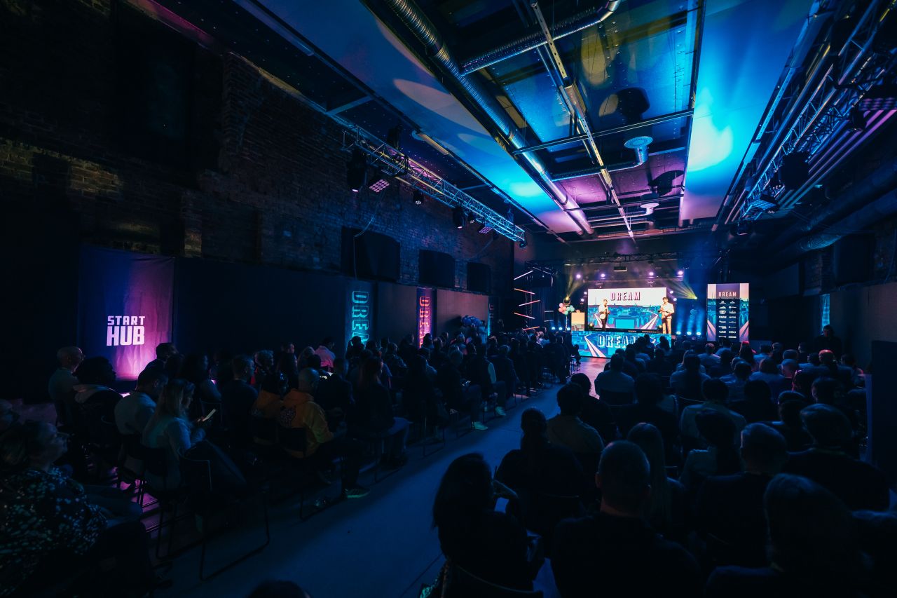 A dark brick hall lit in blue and pink at Dream 2026 in Lahti, with the audience seated in rows facing a stage where two people stand in front of a large Dream screen and a StartHub banner hangs on the left wall.