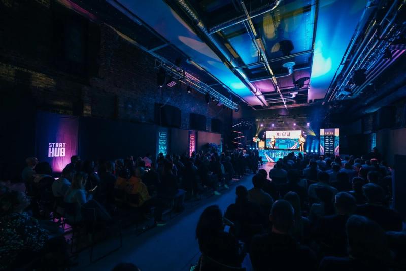 A dark brick hall lit in blue and pink at Dream 2026 in Lahti, with the audience seated in rows facing a stage where two people stand in front of a large Dream screen and a StartHub banner hangs on the left wall.