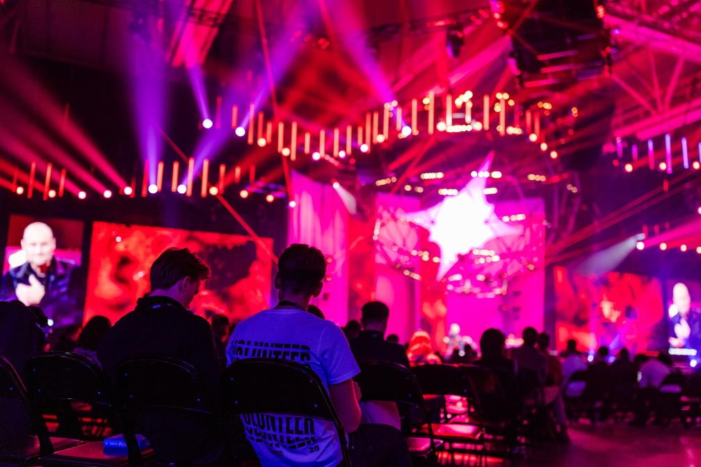 Seen from behind, audience members sit in a hall lit in pink and purple and watch speakers shown on large screens beside the stage.