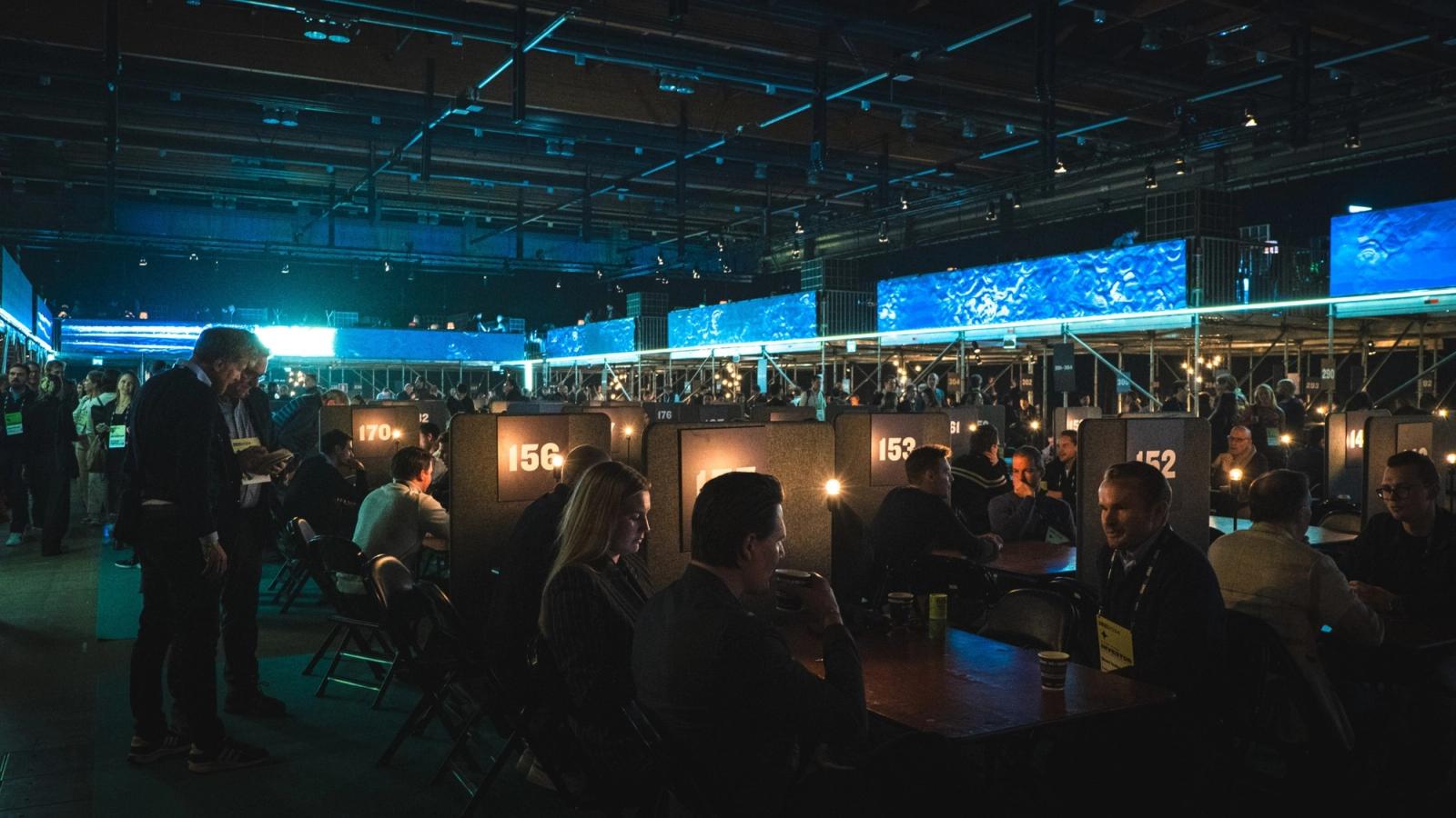 The Slush 2025 meeting area in a dark hall: rows of numbered tables with small lamps, with people seated in pairs and talking, and others walking between the tables.