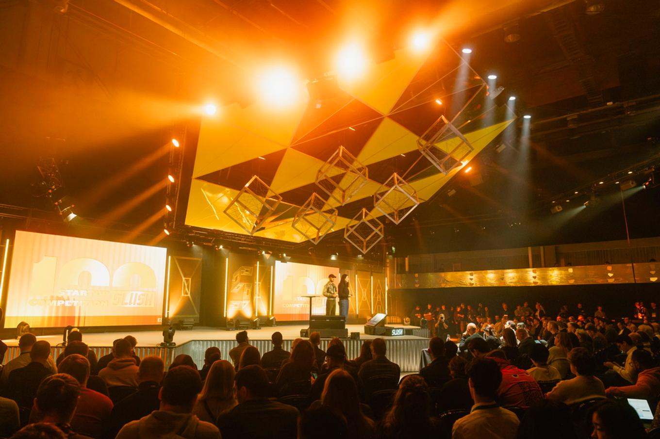 Two presenters stand on a stage under a yellow geometric ceiling installation and orange lights, with a seated audience in the foreground.