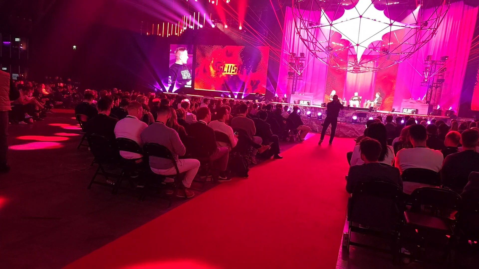 The Founder Stage at Slush 2025 in red and purple light, with people speaking on stage and a seated audience listening.