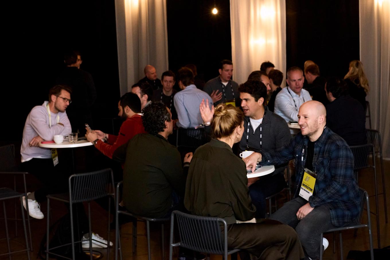 Groups of people sit and stand around small tables in a dim hall, talking in pairs and threes. One man in the foreground wears a yellow Investor badge.