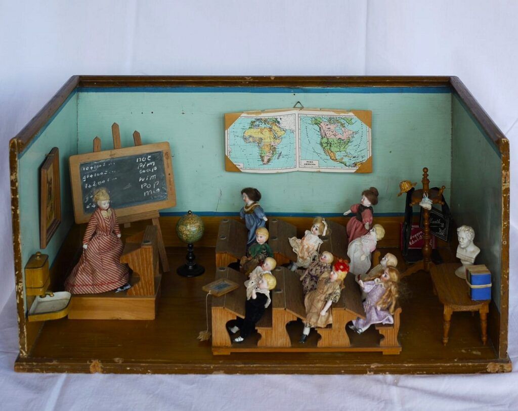 Diorama eines alten Schulzimmers mit Porzellanpuppen in Schulbänken, Wandtafel und Landkarte.