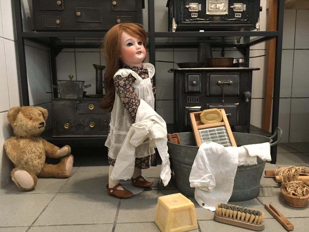 Puppe mit Wäsche vor einem gusseisernen Spielzeugherd, daneben ein alter Teddybär und ein Zinkzuber.
