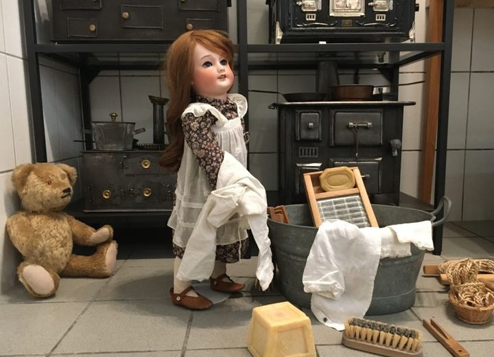 Puppe mit Wäsche vor einem gusseisernen Spielzeugherd, daneben ein alter Teddybär und ein Zinkzuber.