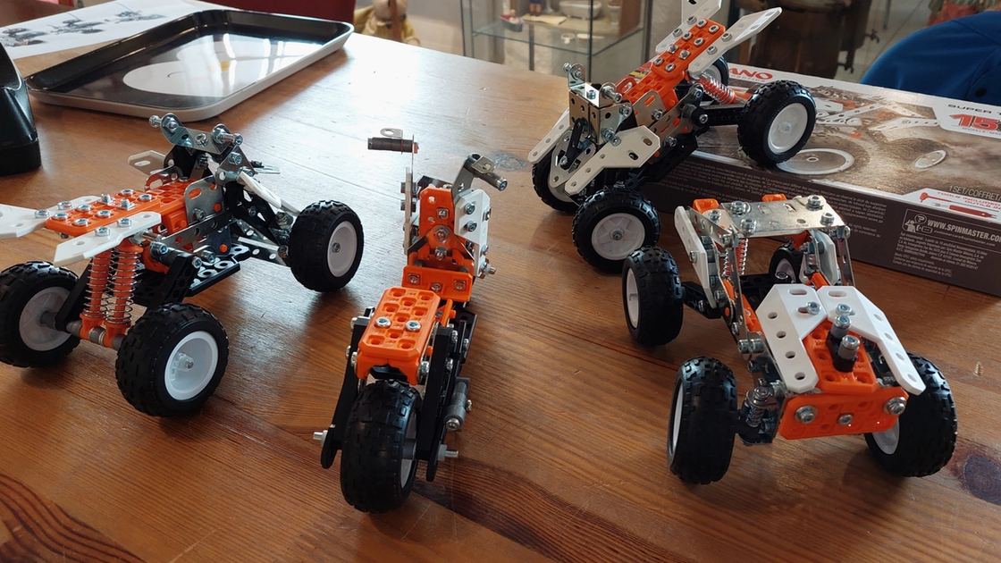 Vier fertige Fahrzeugmodelle aus orangem und silbernem Metallbaukasten auf einem Holztisch