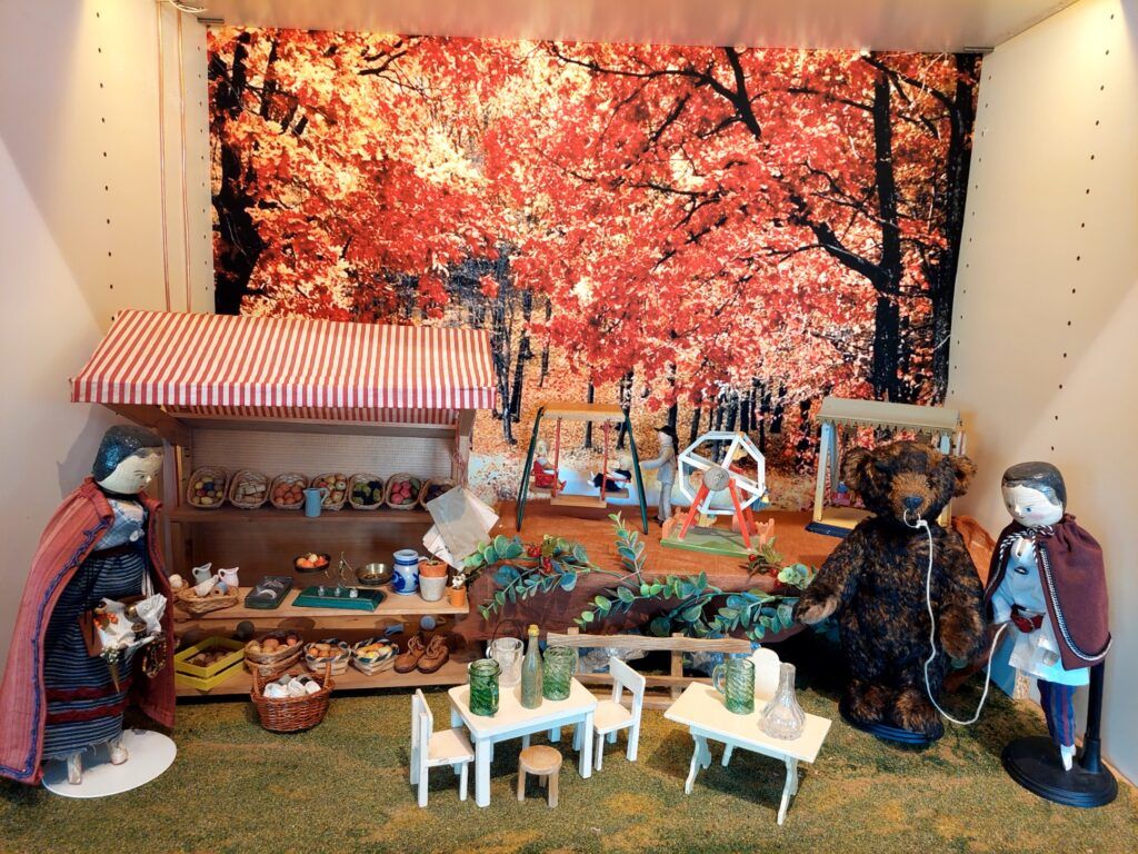 Ausstellungsraum Stadt–Land: Marktstand mit Waren, kleine Tische und Stühle, ein Teddybär und zwei Puppen.