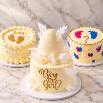 An elegant, cream-colored baby shower cake featuring a soft velvet texture and delicate white edible feathers. A large matching egg ornament sits on top, surrounded by feathers to symbolize a nest. A shimmering gold "Boy or Girl?" topper is displayed on the front, set against a rustic wooden background.