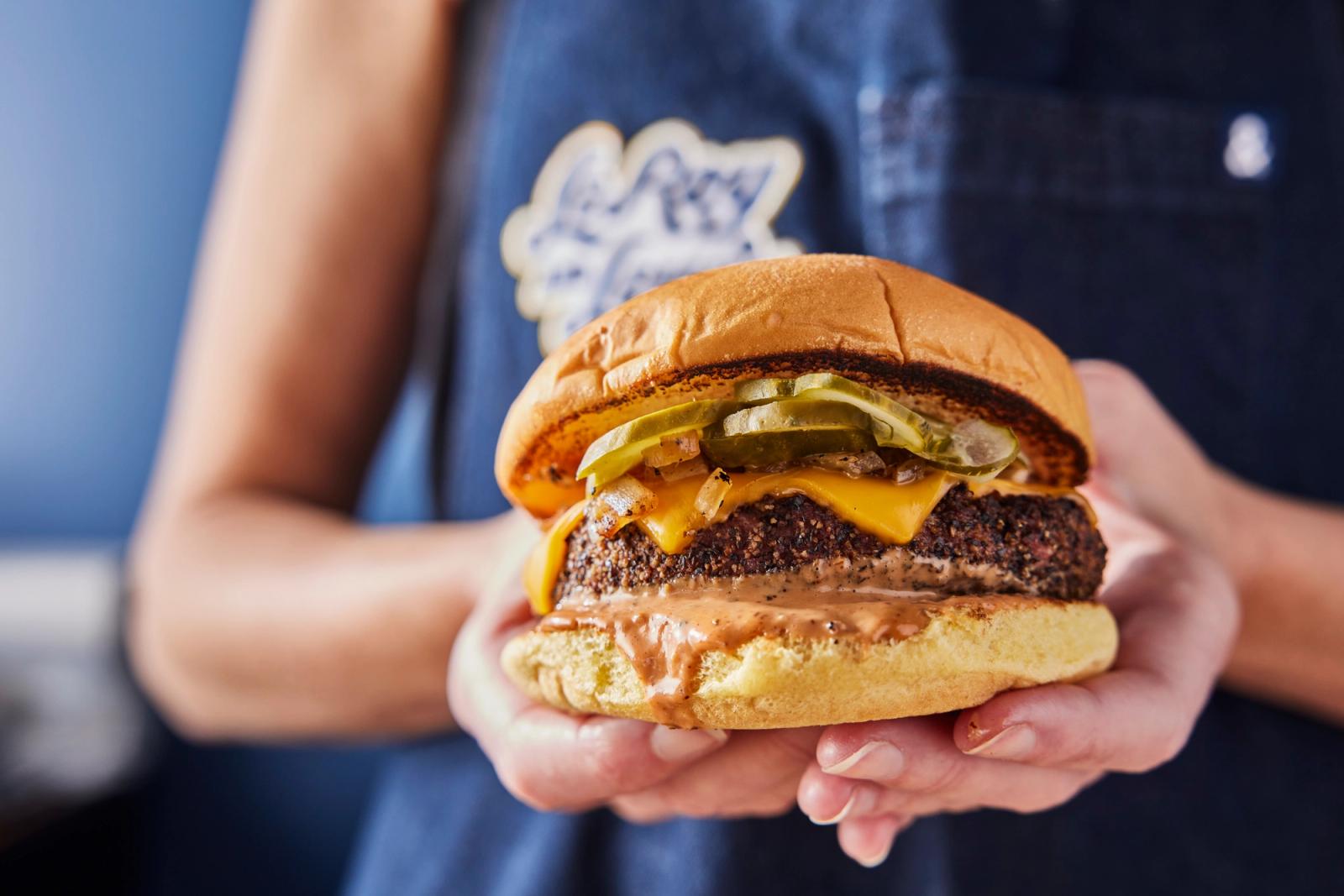 The L&L burger, pickles and melted cheese on a charred bun, held out in two hands in front of a LeRoy and Lewis apron