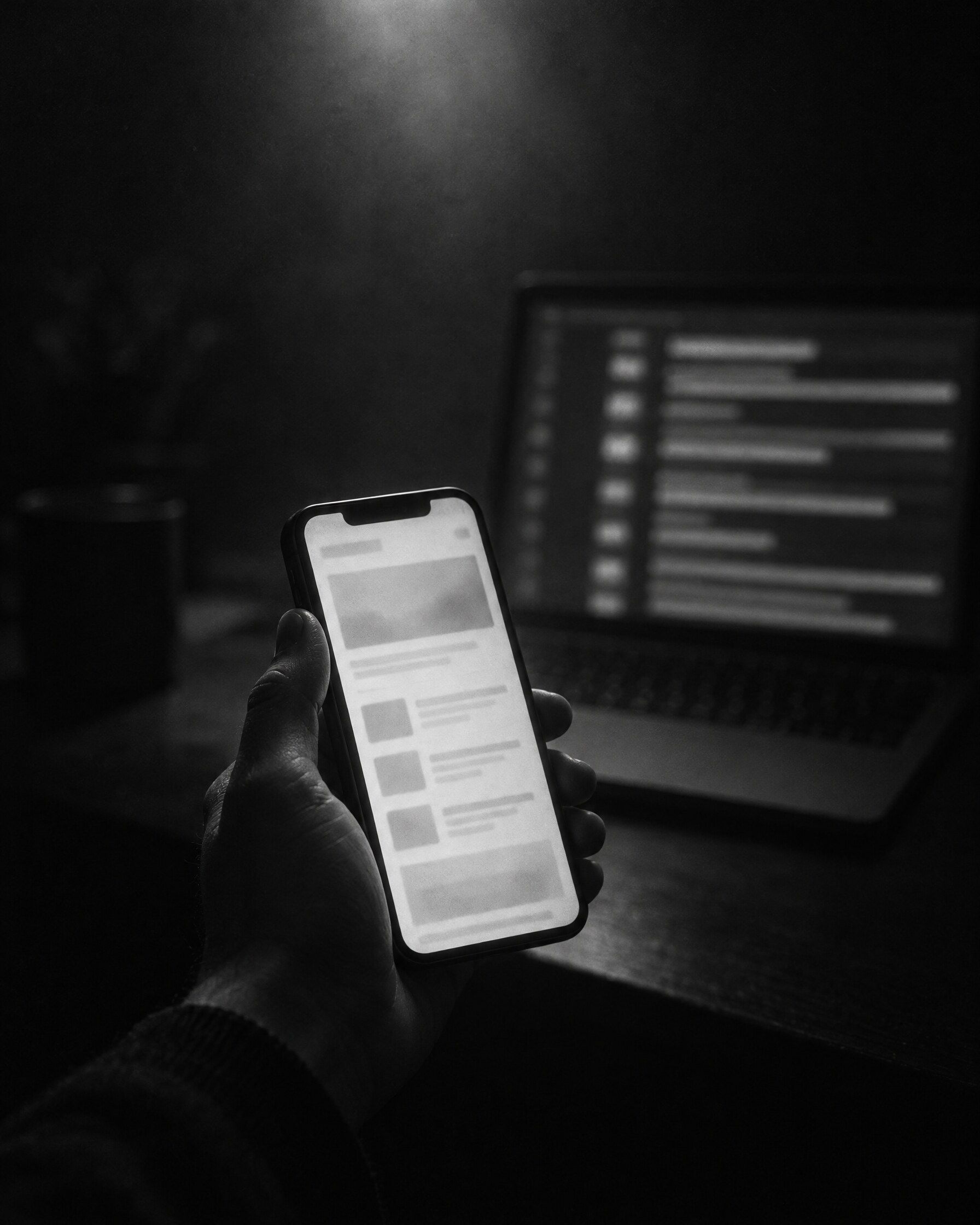 Dłoń trzymająca telefon z rozmytą stroną internetową w ciemnym pokoju, w tle laptop