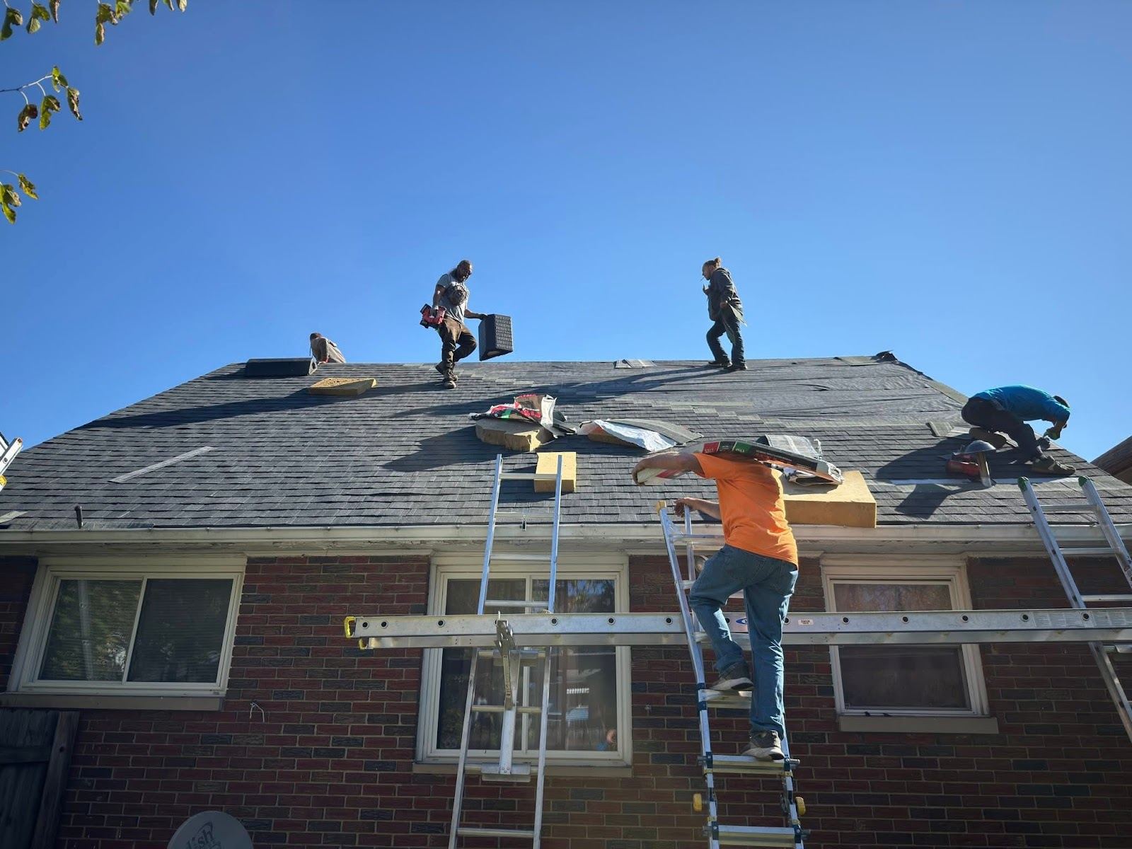 Four roofers on the shingle roof of a brick house, with ladders up the front wall