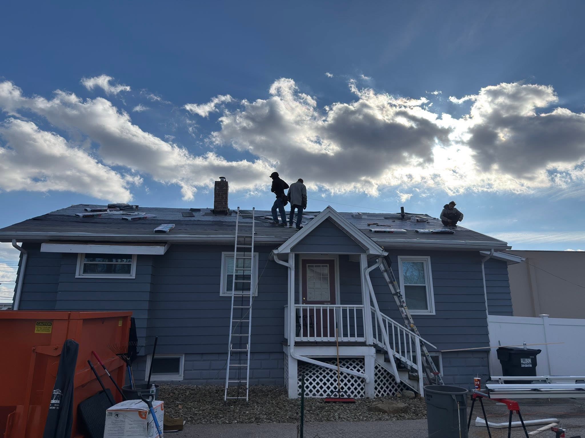 A crew of three on the roof of a blue house, ladders up and a dumpster in the yard