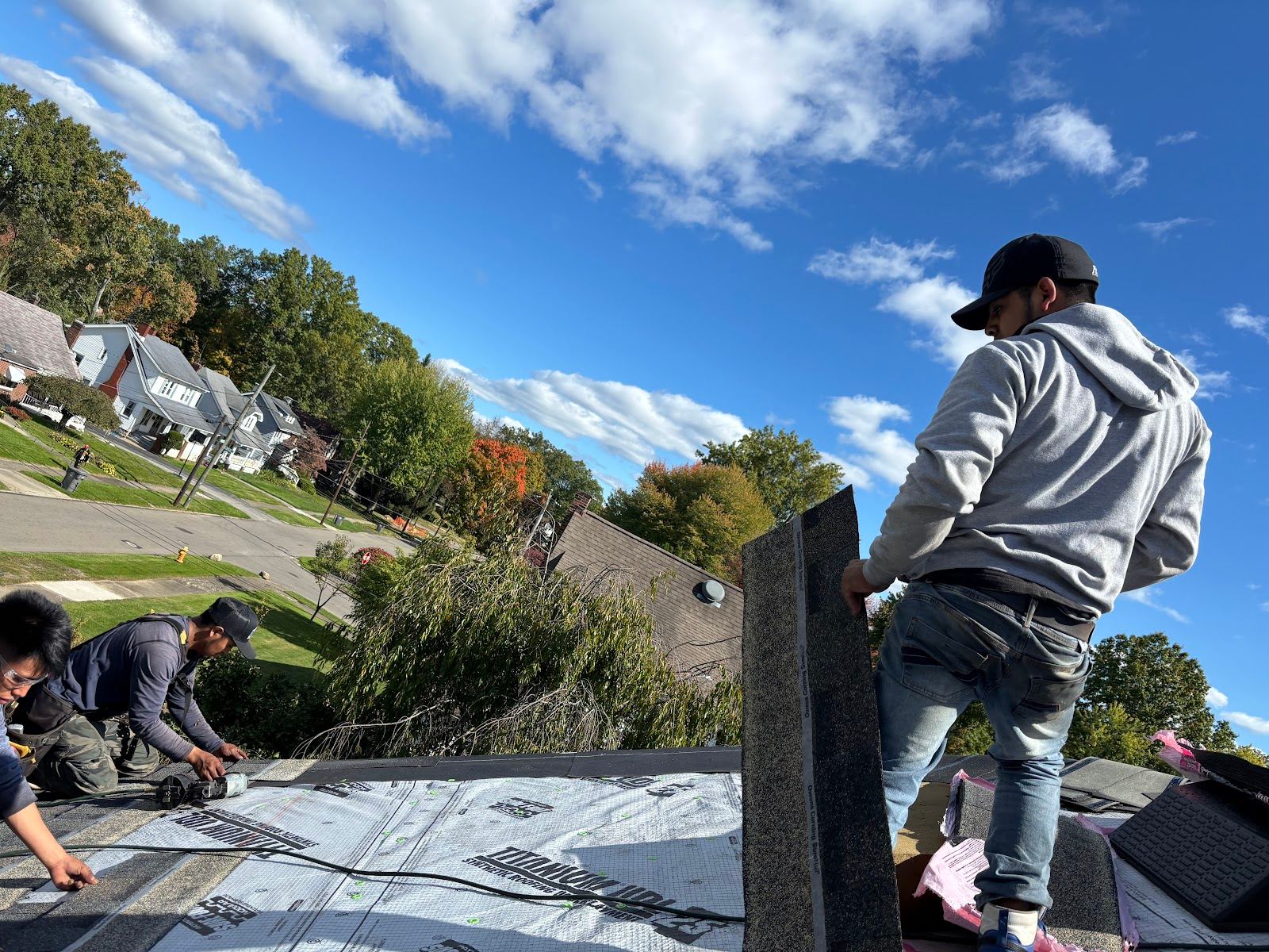 Roofers at work on a roof above a residential street, one carrying a bundle of shingles