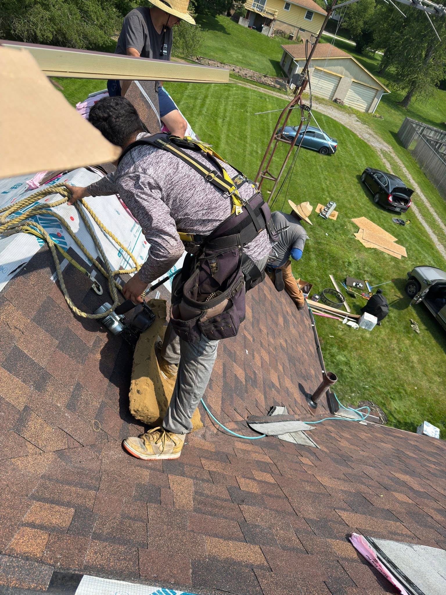 A roofer in a harness nailing brown shingles near the edge of a roof