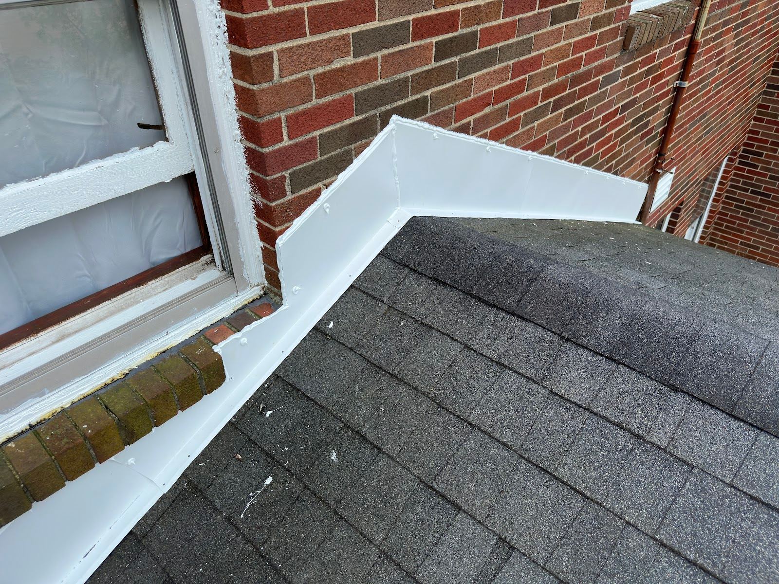 New white metal flashing where a shingle roof meets a brick wall, under a window