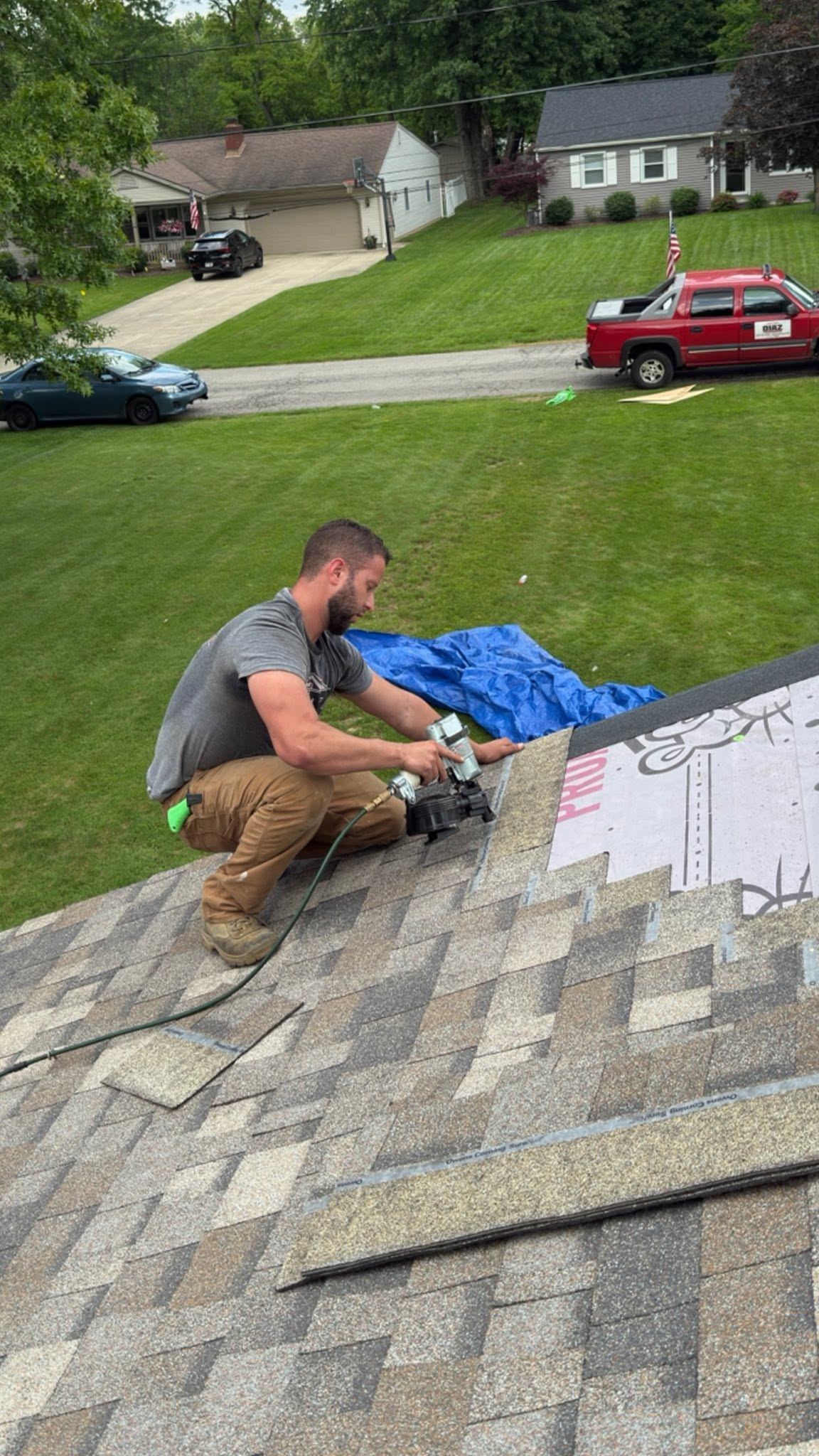 A roofer cutting shingles on a roof, a blue tarp spread out behind him