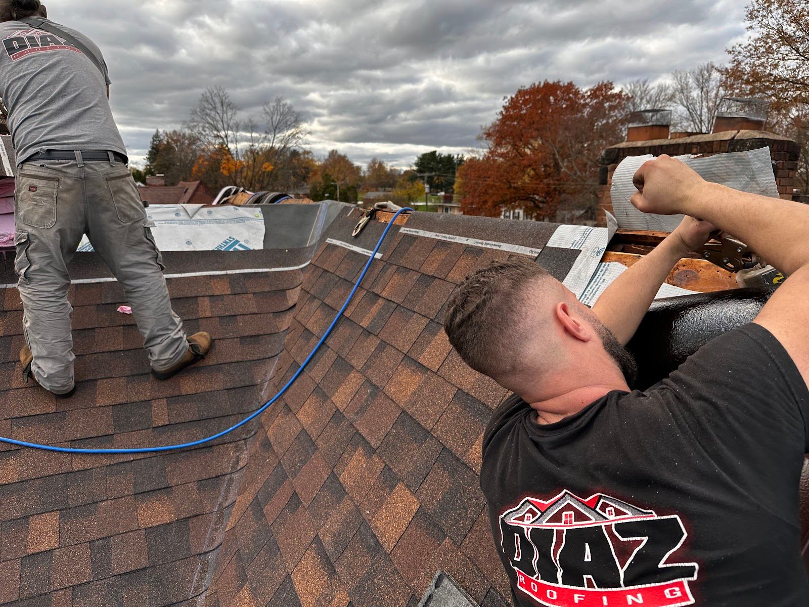 A Diaz Roofing roofer in a branded shirt working at a chimney while a teammate works the slope behind him