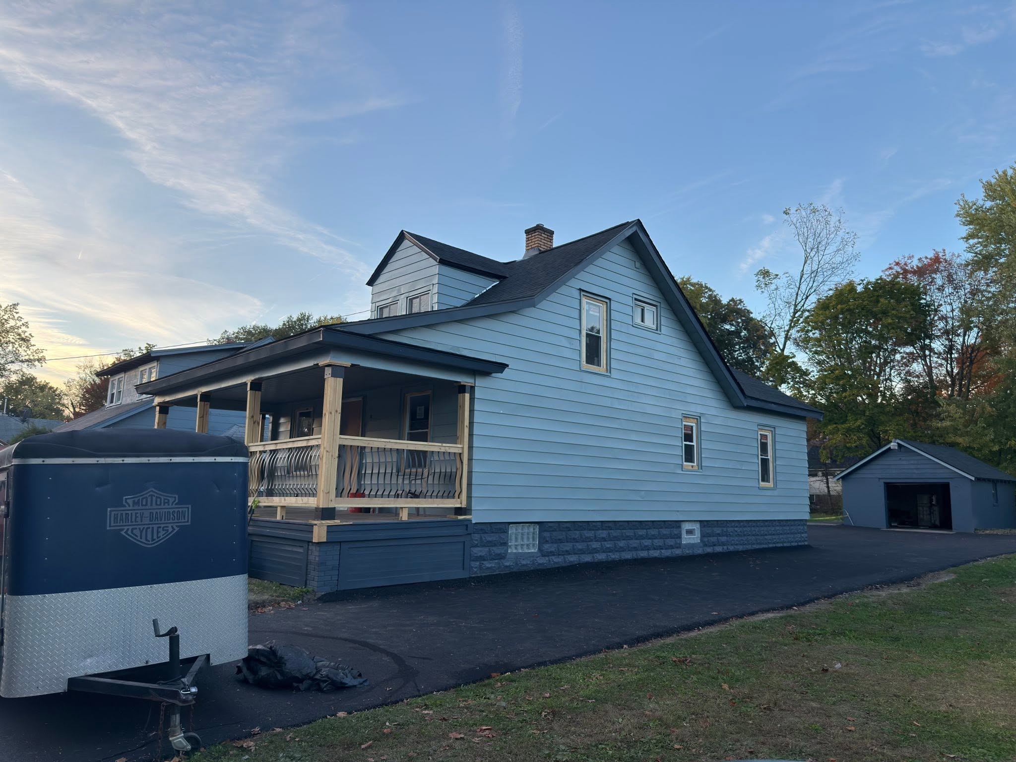 A blue house with a dark shingle roof and the work trailer beside it