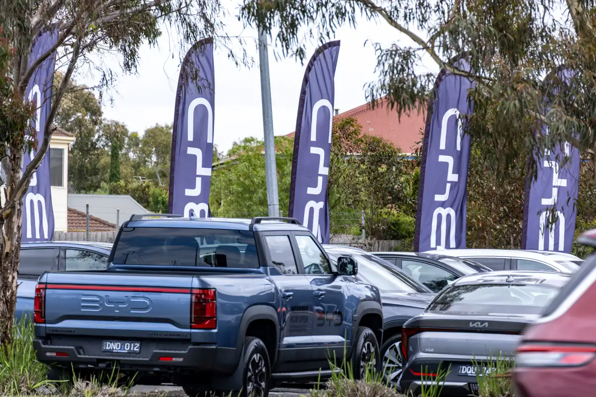 BYD logo on branded flags at an outdoor car dealership with a lineup of parked BYD electric vehicles in suburban Australia. BYD is a major Chinese multinational automaker known for electric vehicles