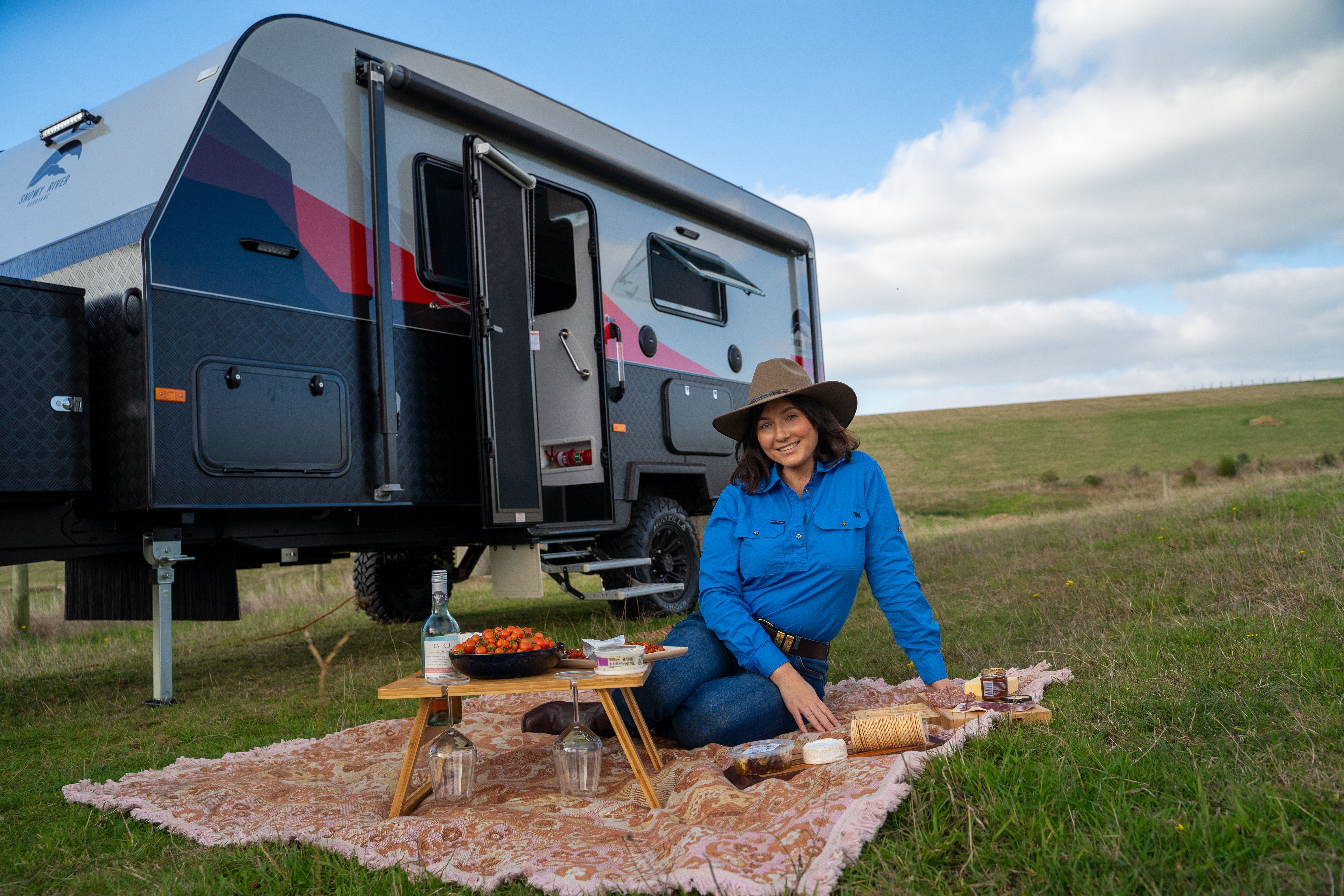 woman in blue shirt on a rug  having a picnic in a field in front of a caravan