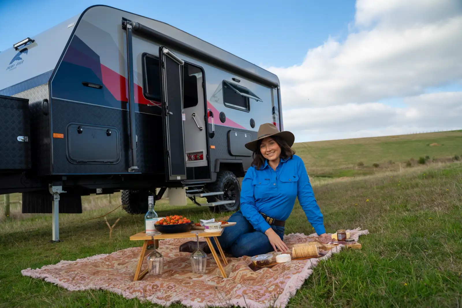 woman in blue shirt on a rug having a picnic in a field in front of a caravan