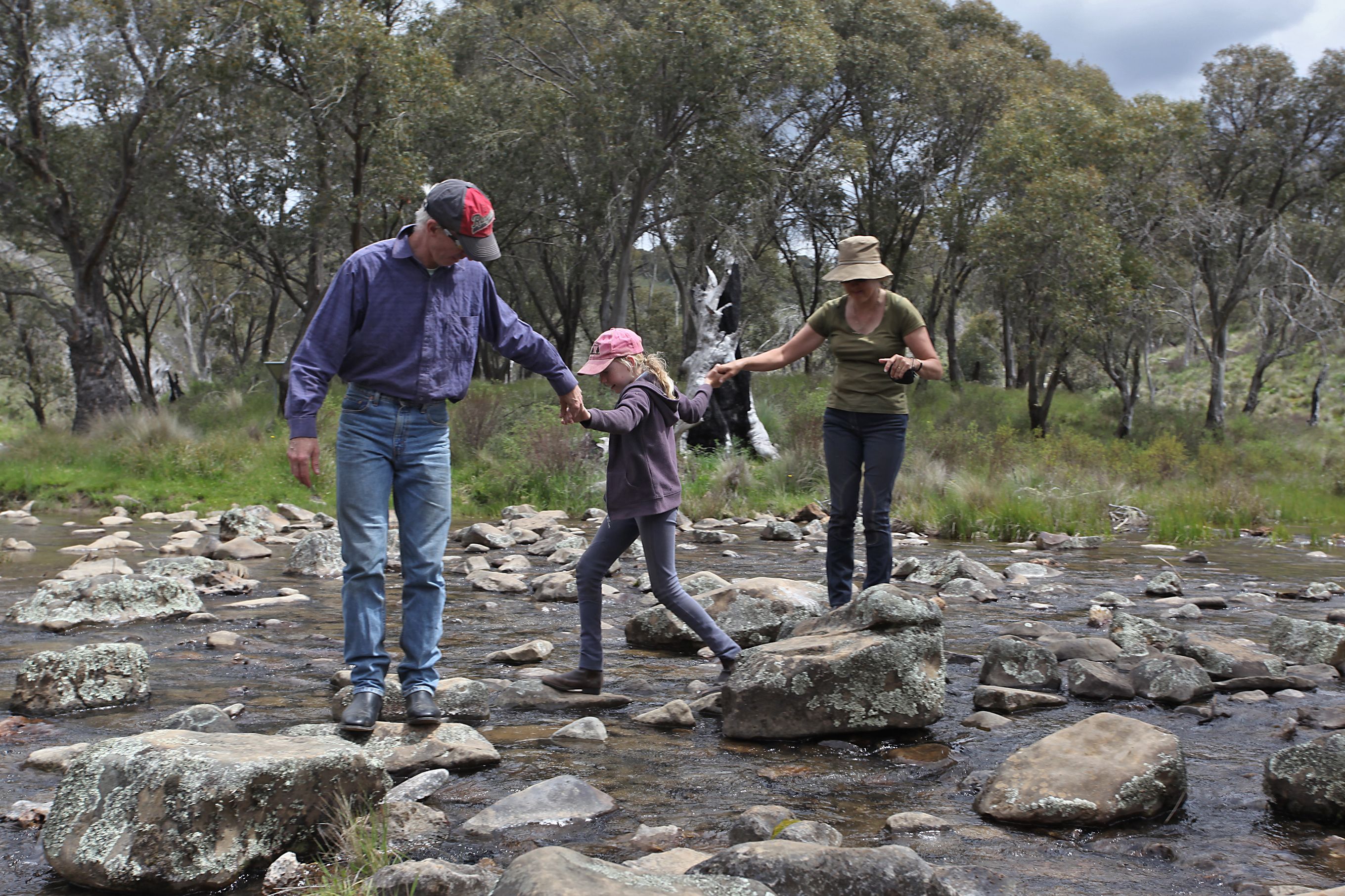 Two adults and a girl holding hands as they hop across a river on rocks