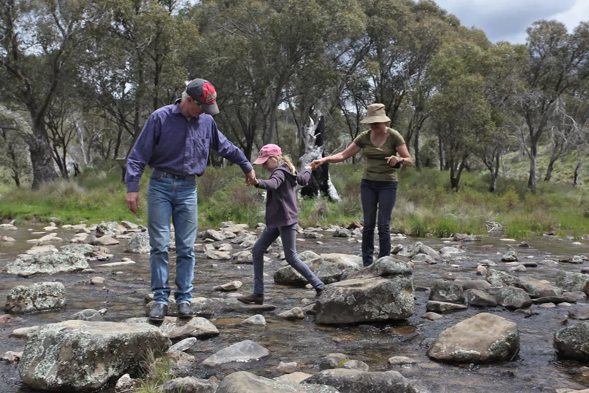 Two adults and a girl holding hands as they hop across a river on rocks
