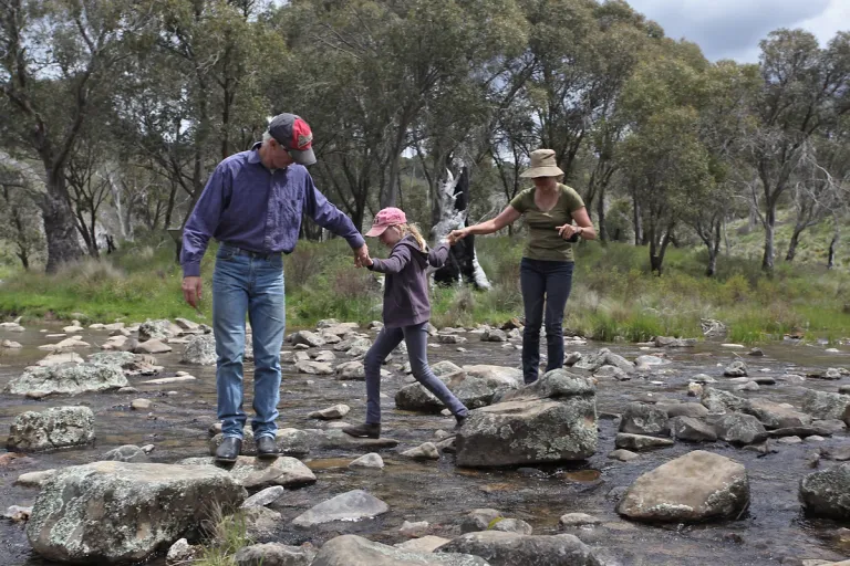 Two adults and a girl holding hands as they hop across a river on rocks