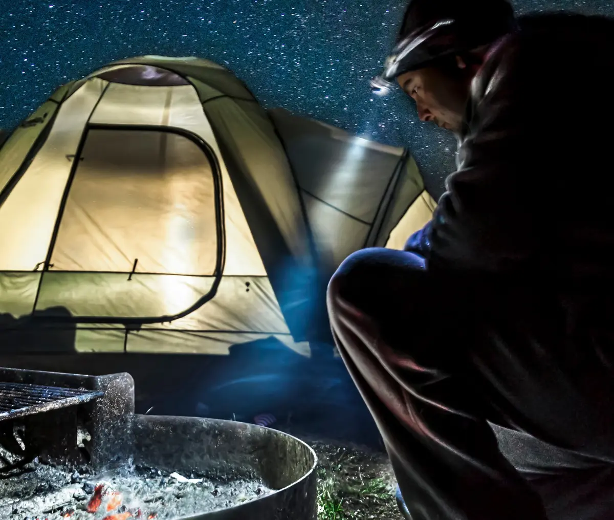 man sat over a campfire with his head torch shining and a tent in the backgorund