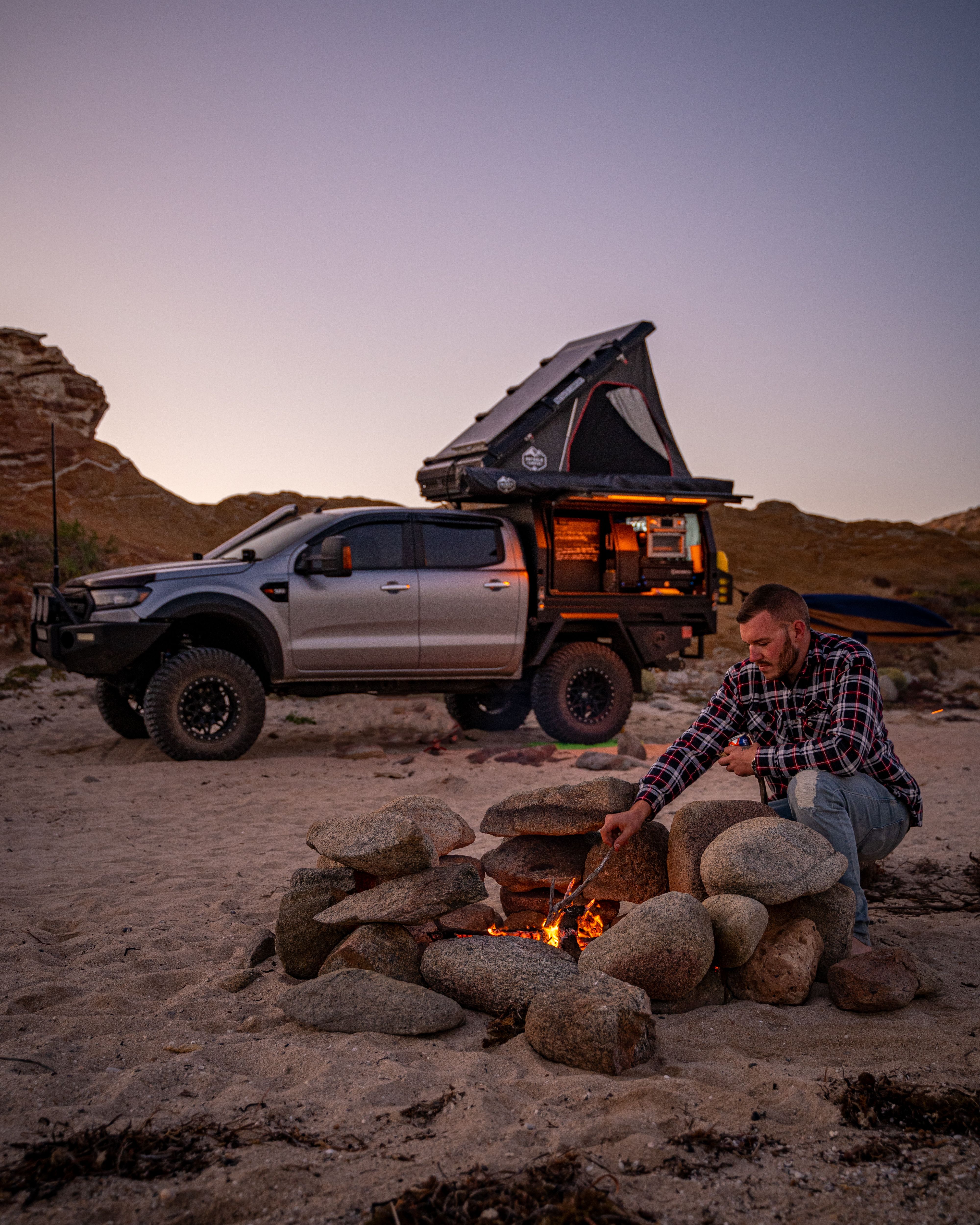 Josh Leonard stoking a campfire with his 4X4 Ford Ranger in the background in Australia's Outback during a sunset