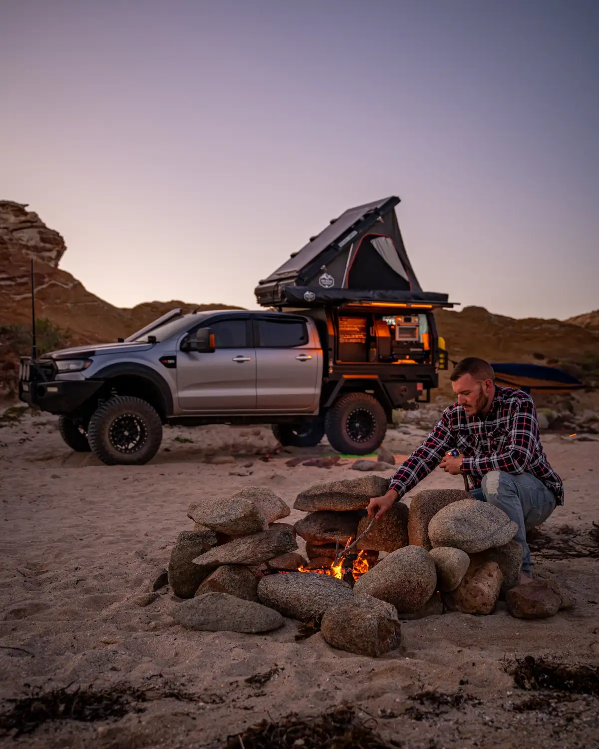 Josh Leonard stoking a campfire with his 4X4 Ford Ranger in the background in Australia's Outback during a sunset
