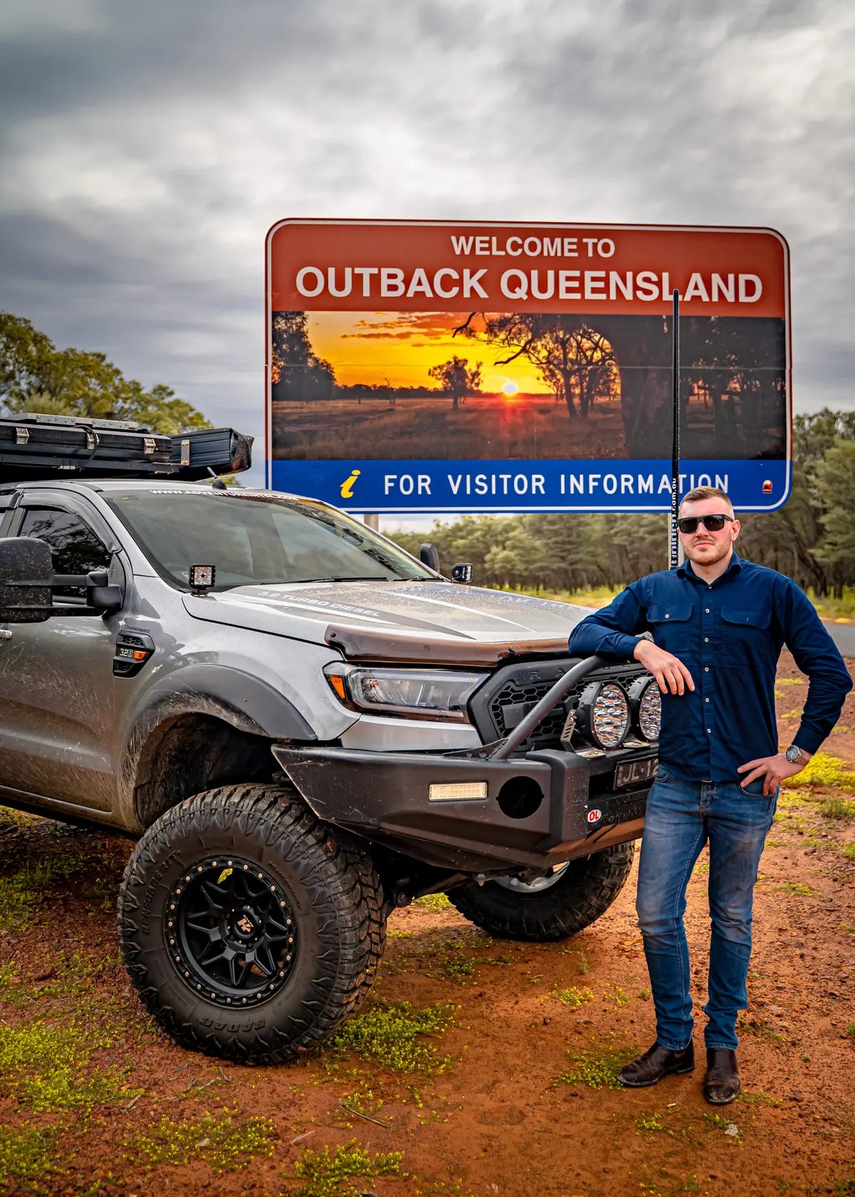 A man posing with his 4X4 Ford Ranger in front of a welcome to Outback Queensland road sign