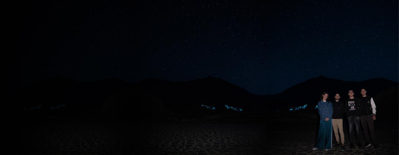 A small group standing together under a starry night sky, mountains behind them