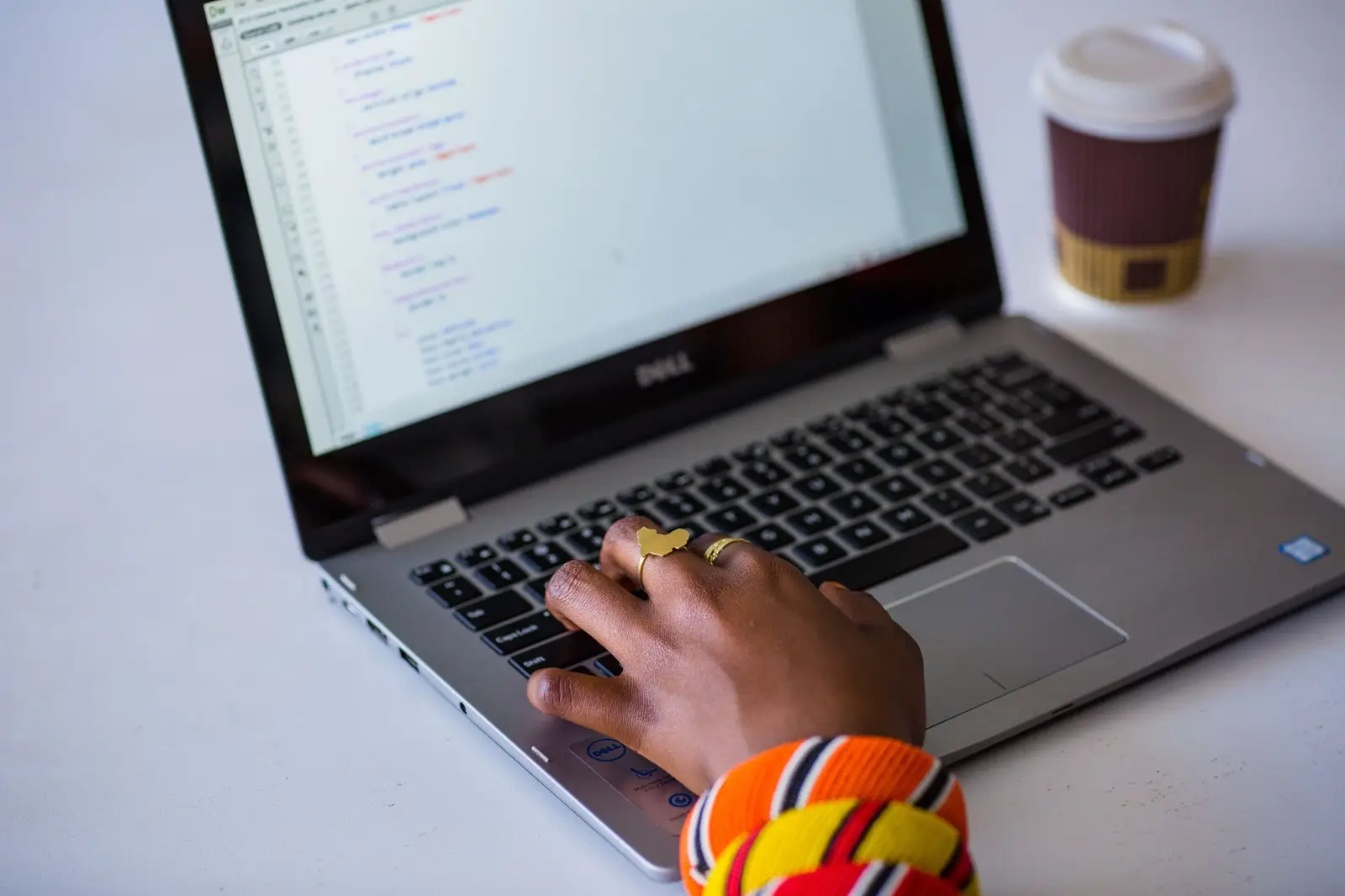 Black person's hand wearing two gold rings, typing on a laptop next to a to-go cup of coffee