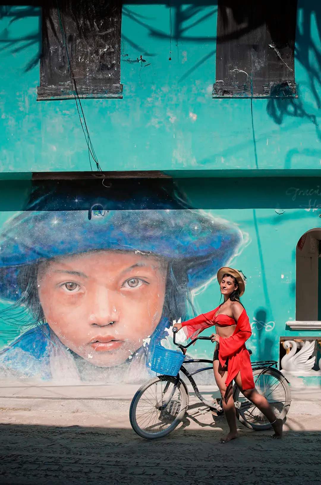 Woman in red outfit and hat posing with a bicycle in front of a mural of a child's face