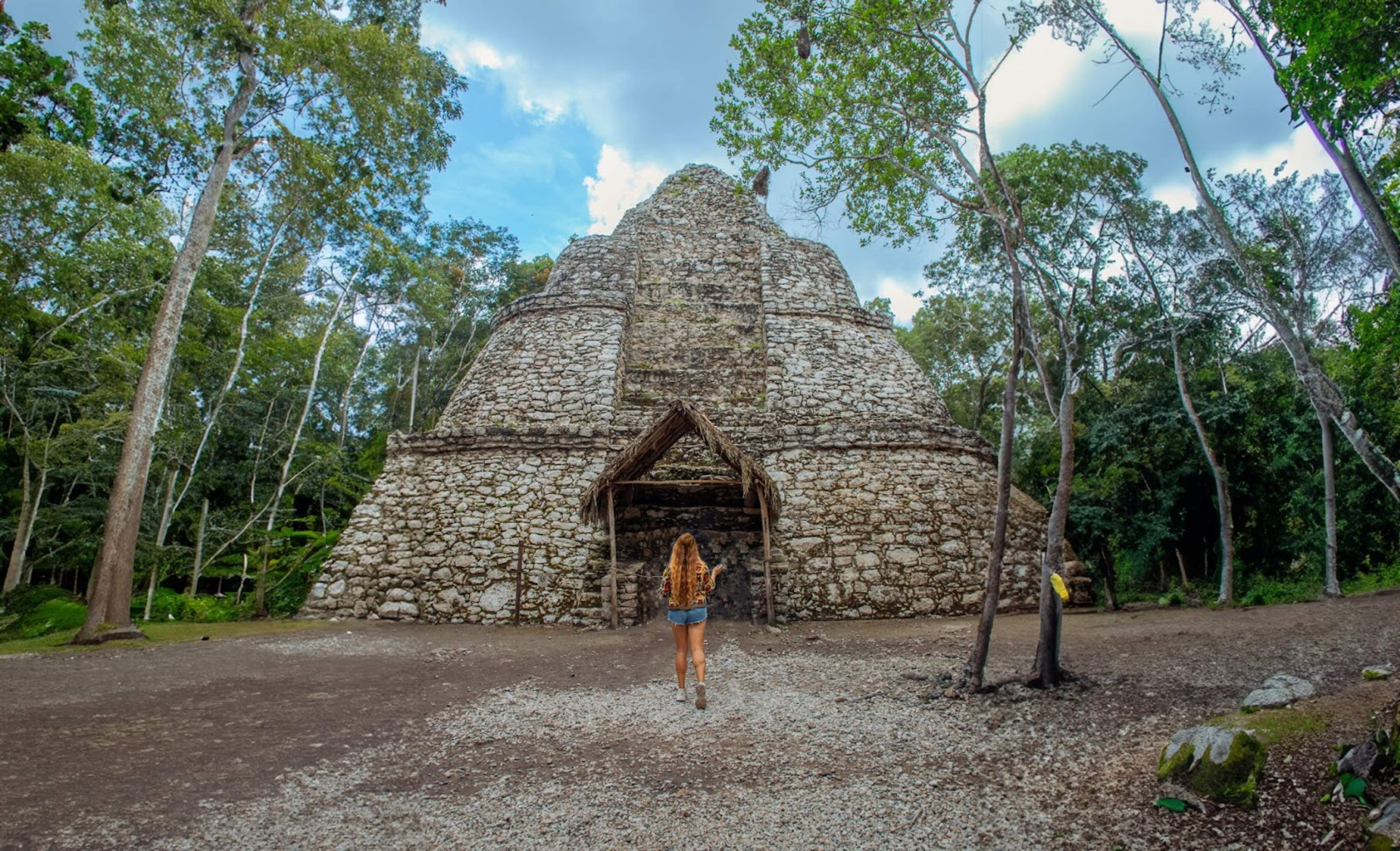 A woman with long hair walks toward a large, ancient Mayan pyramid surrounded by dense forest and trees.