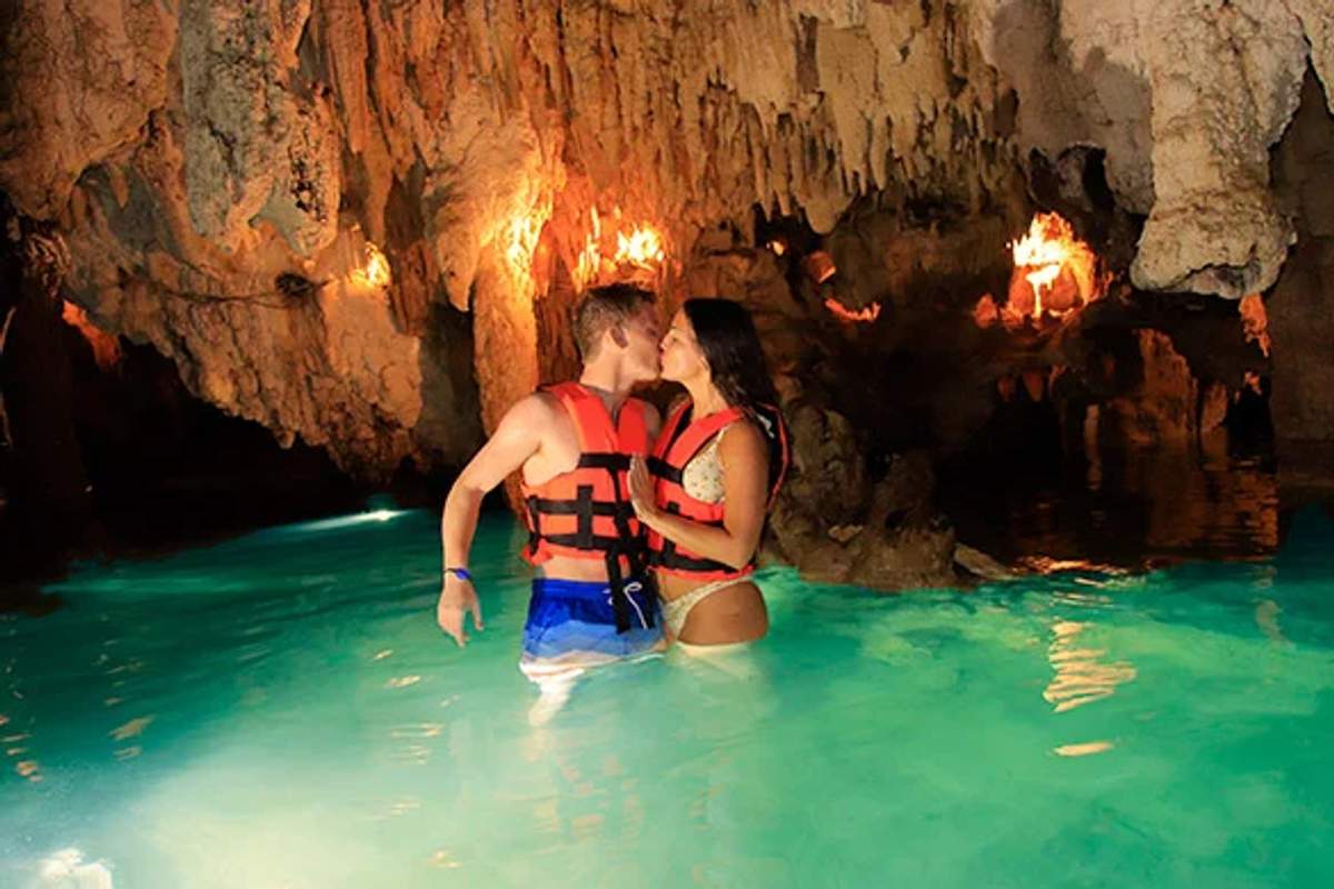 A couple wearing life vests shares a kiss while standing in the illuminated turquoise waters of a cenote in Cancun.