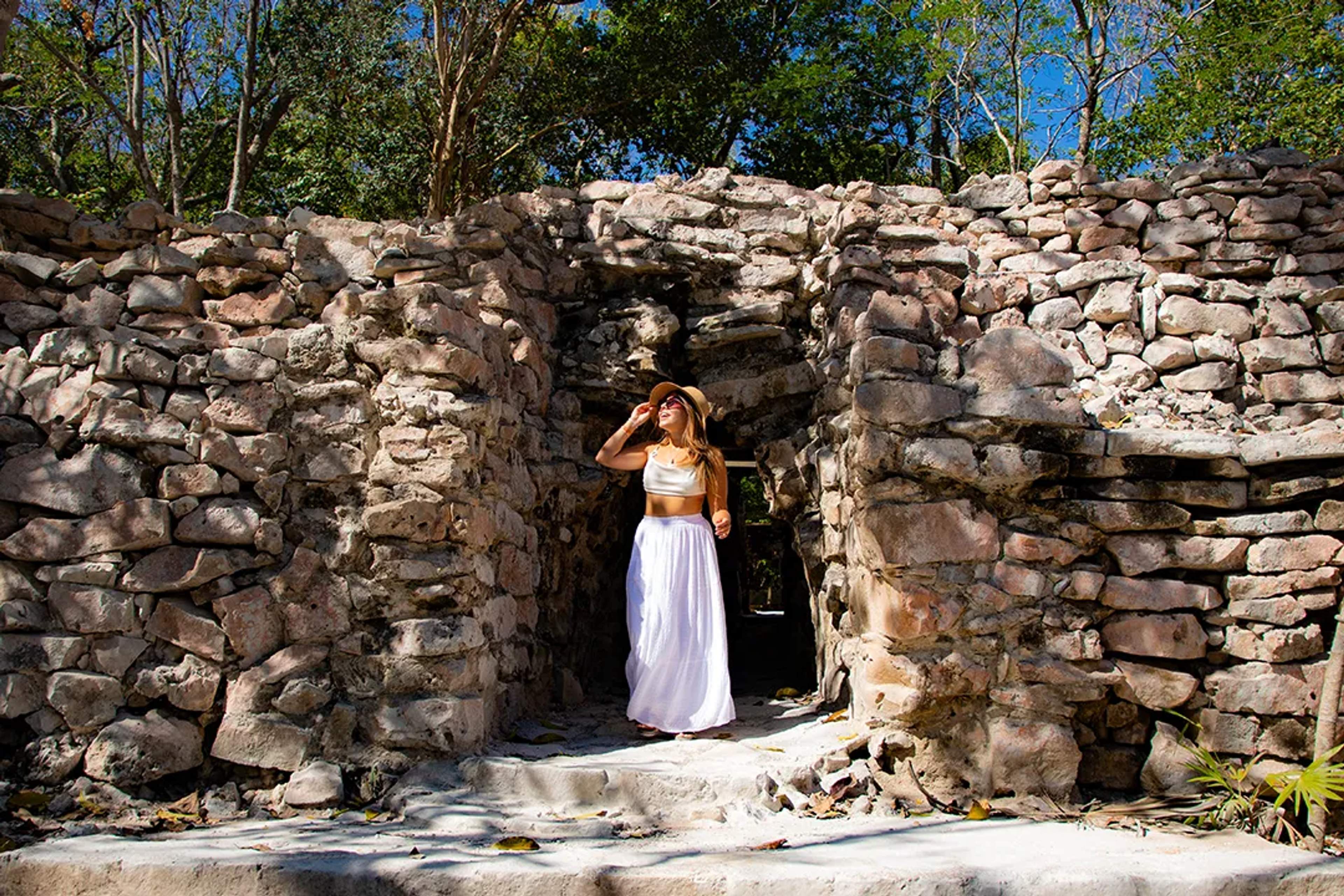 Mujer explorando ruinas de piedra antiguas en la selva, vestida de blanco con sombrero.