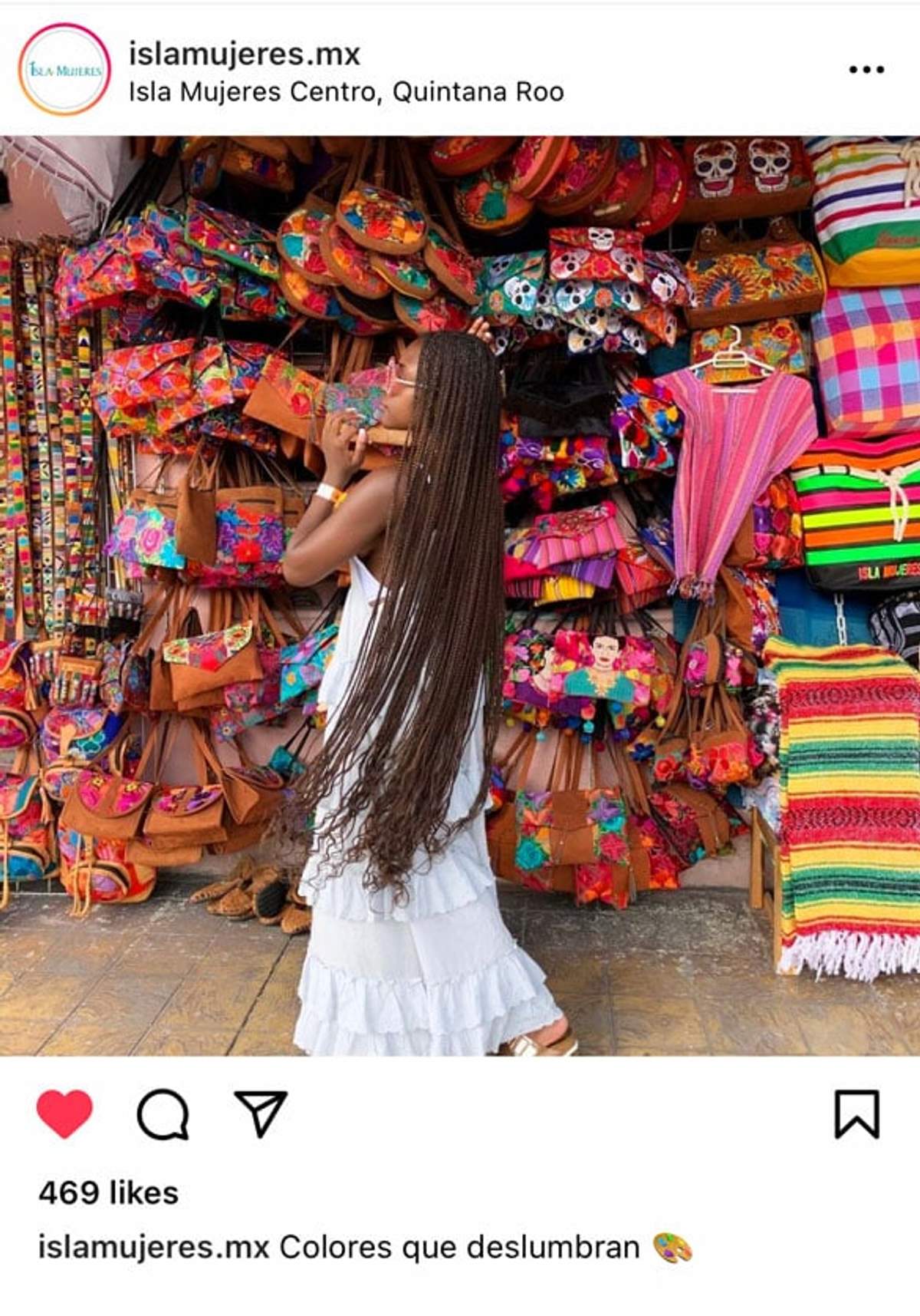 Una mujer con trenzas largas y un vestido blanco comprando en un puesto de mercado colorido lleno de bolsas vibrantes, textiles y artesanías en Isla Mujeres.