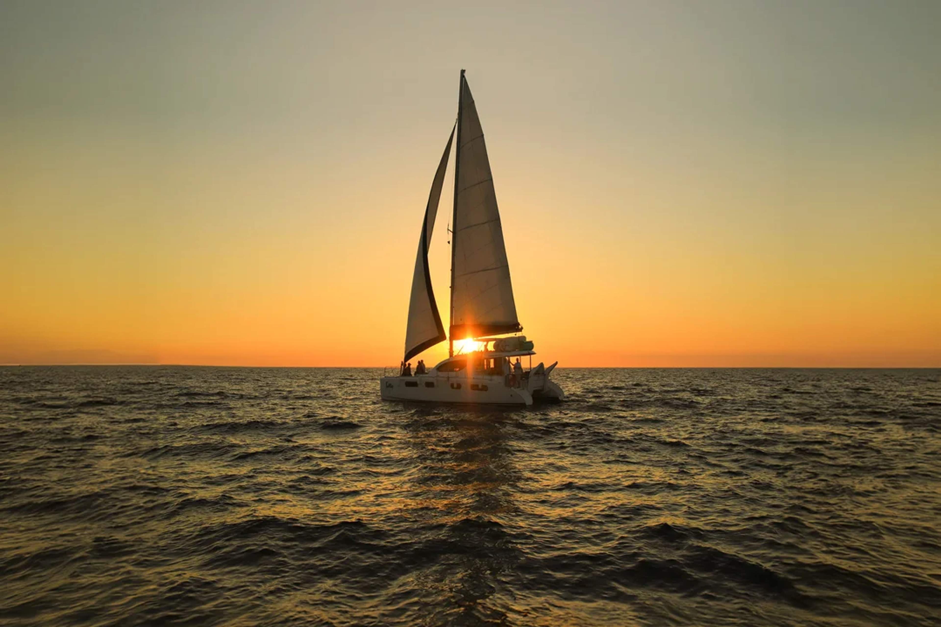 Catamaran sails at sunset on calm waters, with the sun setting behind its sails.