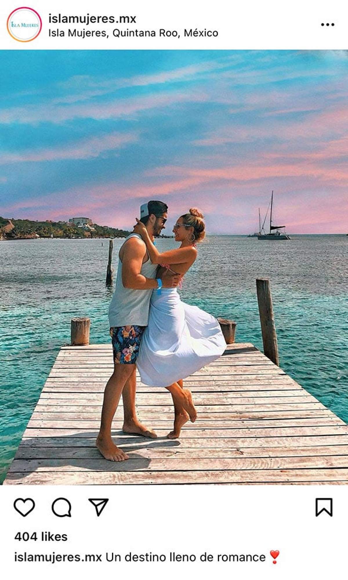 Una pareja se abraza en un muelle de madera sobre el agua turquesa con un velero al fondo y un cielo colorido al atardecer en Isla Mujeres.