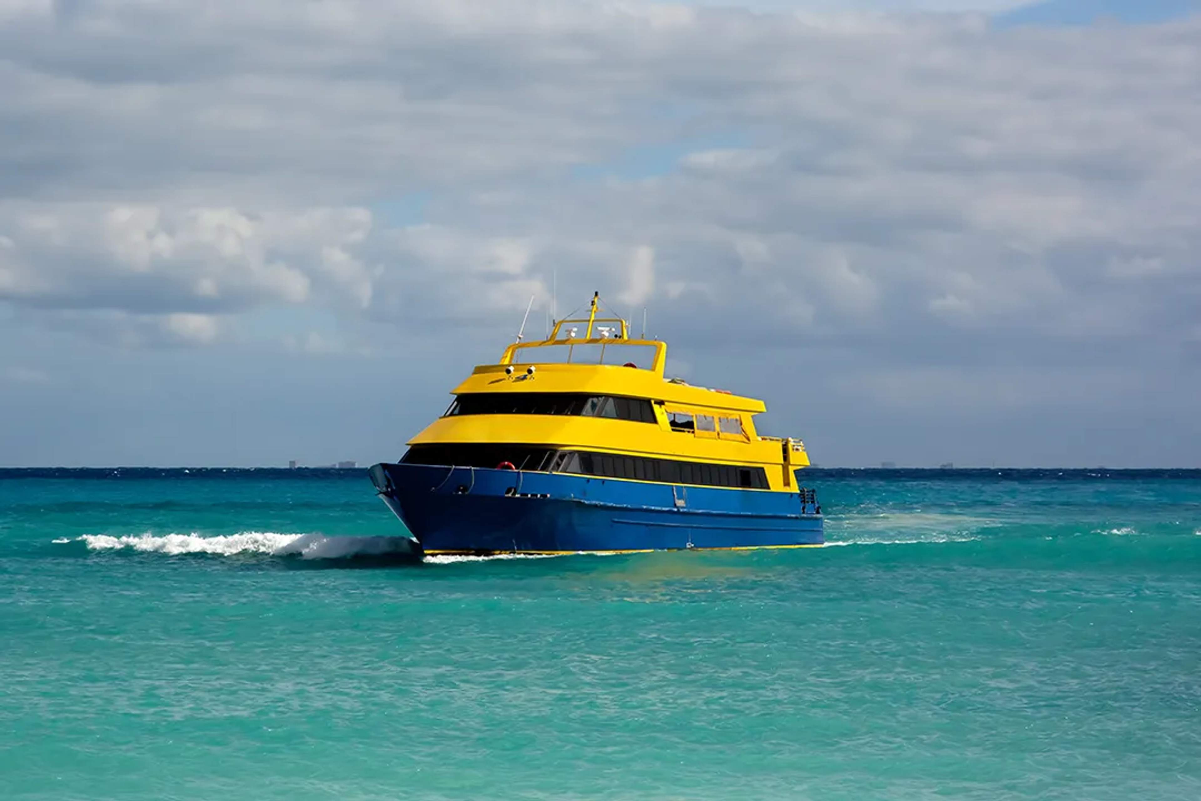 Bright yellow and blue ferry cruising through turquoise Caribbean waters under cloudy skies.