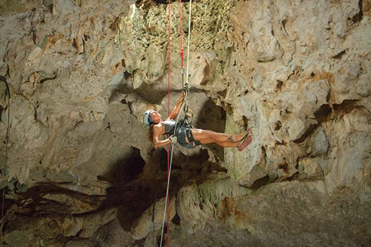 Una mujer haciendo rappel en una cueva en Cancún, equipada con equipo de seguridad y rodeada de paredes rocosas.