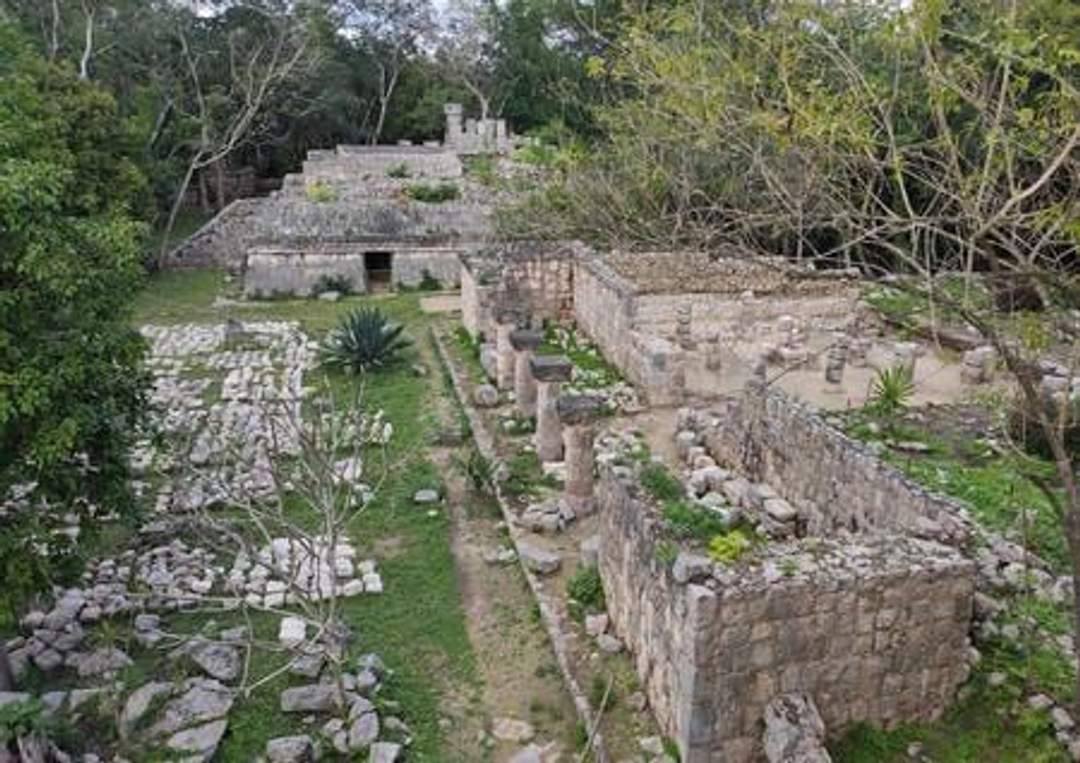 A photograph of the Temple of the Owls at Chichen Itza, featuring an ancient stone structure with multiple columns and a staircase leading to the entrance. The temple is surrounded by lush greenery. Photo credit: Giovanni Agostino Frassetto.
